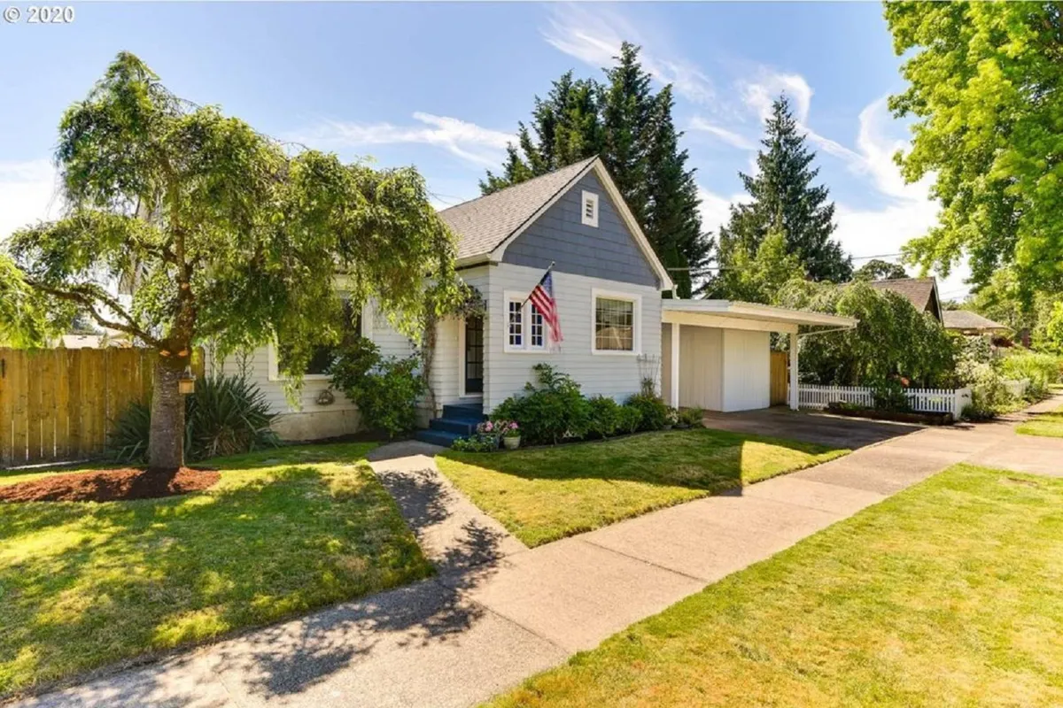 The image shows the charming front exterior of a single-family house in Newberg, Oregon, featuring a classic front porch adorned with an American flag, surrounded by a lush, manicured lawn. The inviting porch suggests a perfect spot for relaxing evenings, while the well-kept yard hints at outdoor family activities and gatherings.
