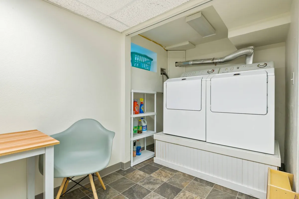 This image displays a practical laundry area featuring a washer and dryer, a shelf neatly organized with cleaning supplies, and a chair. The scene is clean and functional, emphasizing convenience for extended stays.
