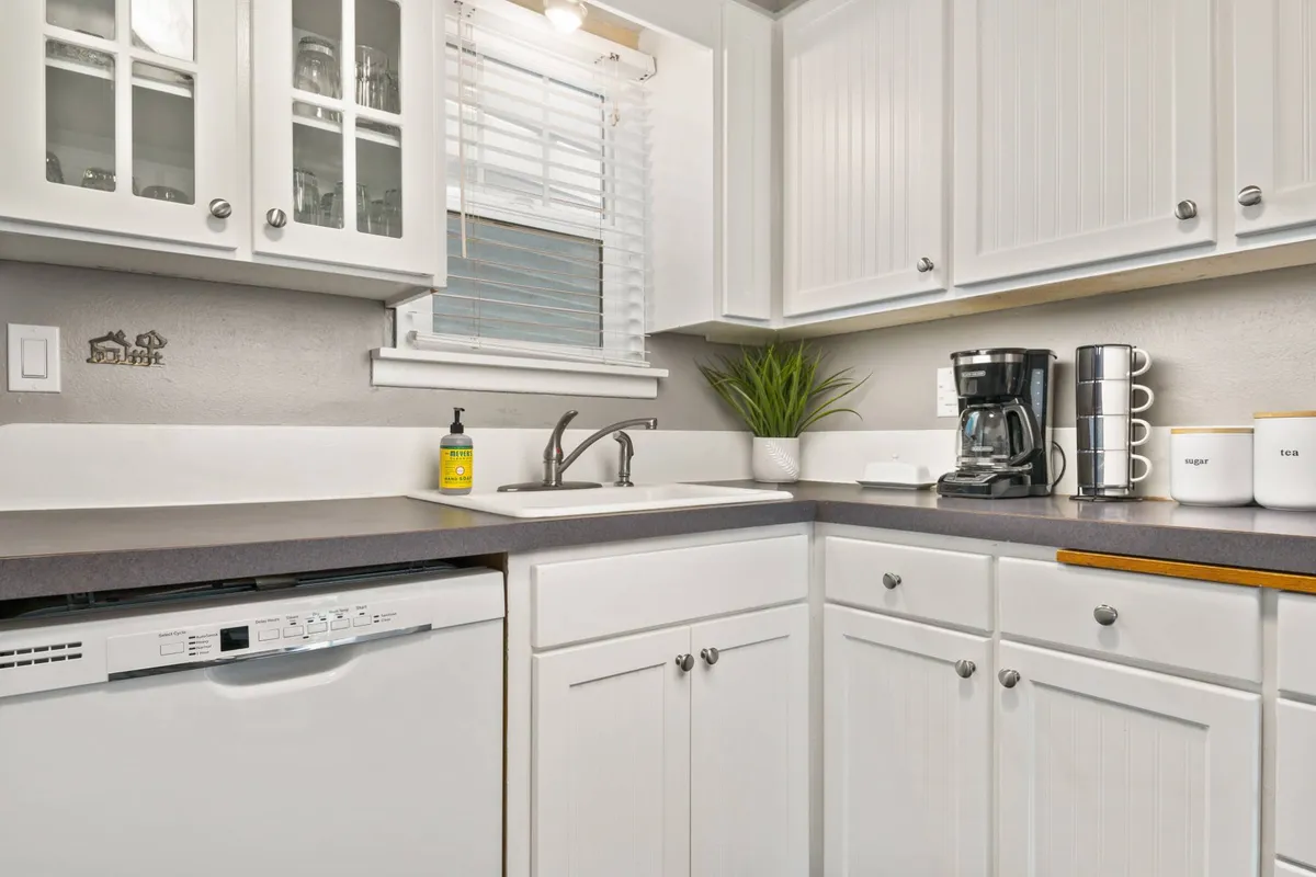 A bright and inviting kitchen scene with a prominent sink, modern coffee maker, a lush green plant, dishwasher, and ample countertop space. The area feels warm and practical, ideal for preparing meals and socializing.