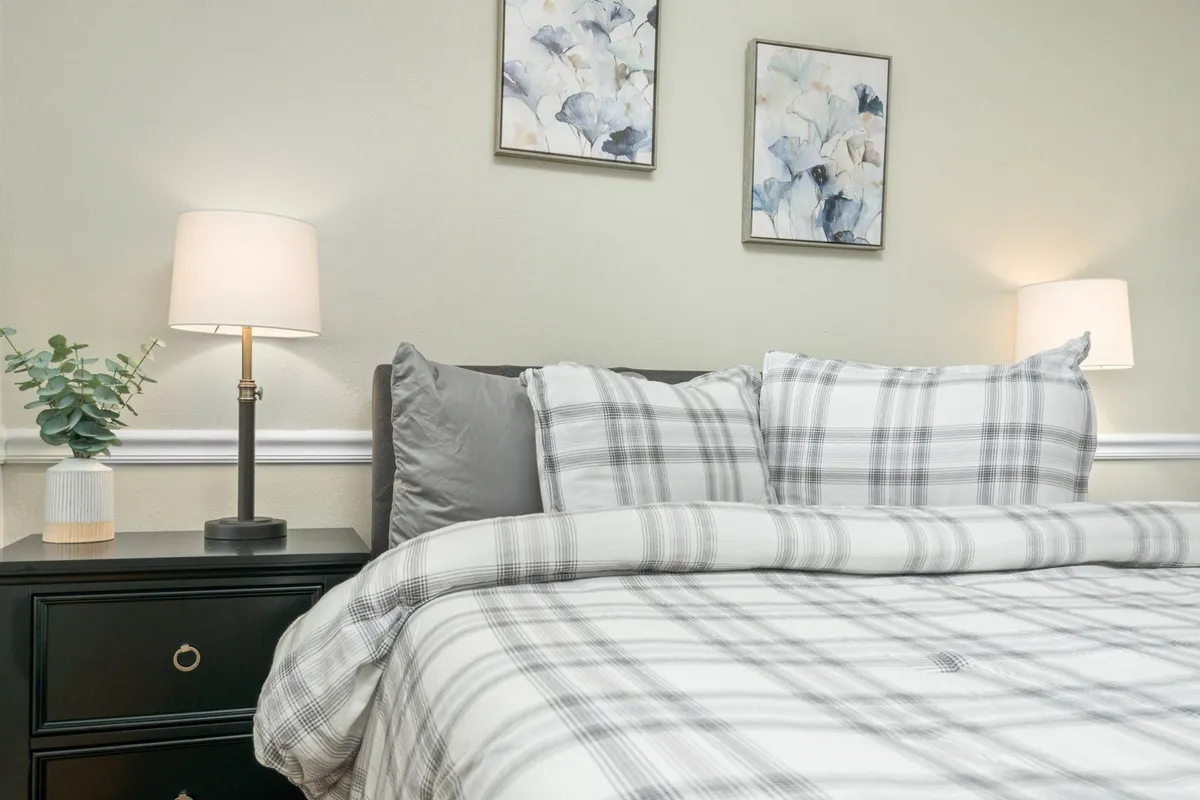 A cozy bedroom featuring a queen bed with plaid bedding, decorative wall art, and a nightstand with a softly glowing lamp. The décor is tranquil, creating a warm and welcoming ambiance.